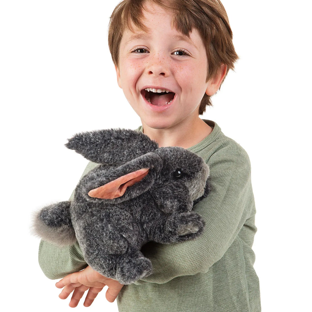 A joyful child in a green shirt holds a plush gray rabbit with floppy ears, smiling widely. The mood is playful and happy, suggesting delight.