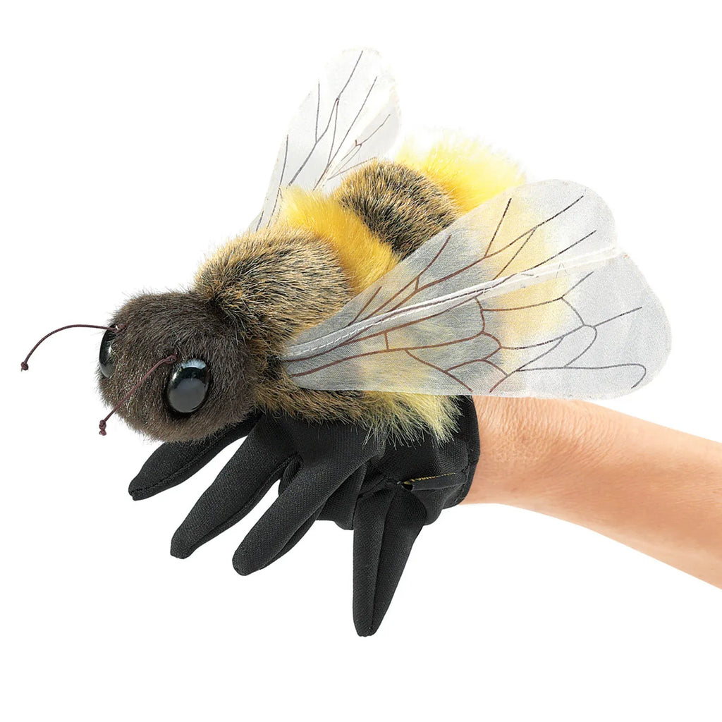 A person wearing a black glove holds a realistic plush bee. The bee has detailed wings, fuzzy yellow and black body, large eyes, and conveys a whimsical tone.