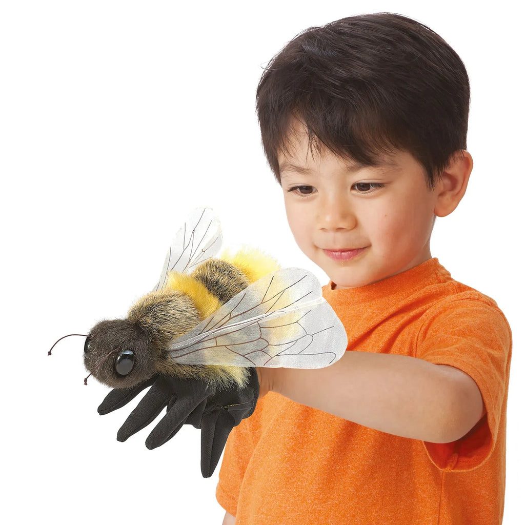 A young child in an orange shirt smiles while holding a large, realistic bee puppet on their hand, conveying curiosity and wonder.