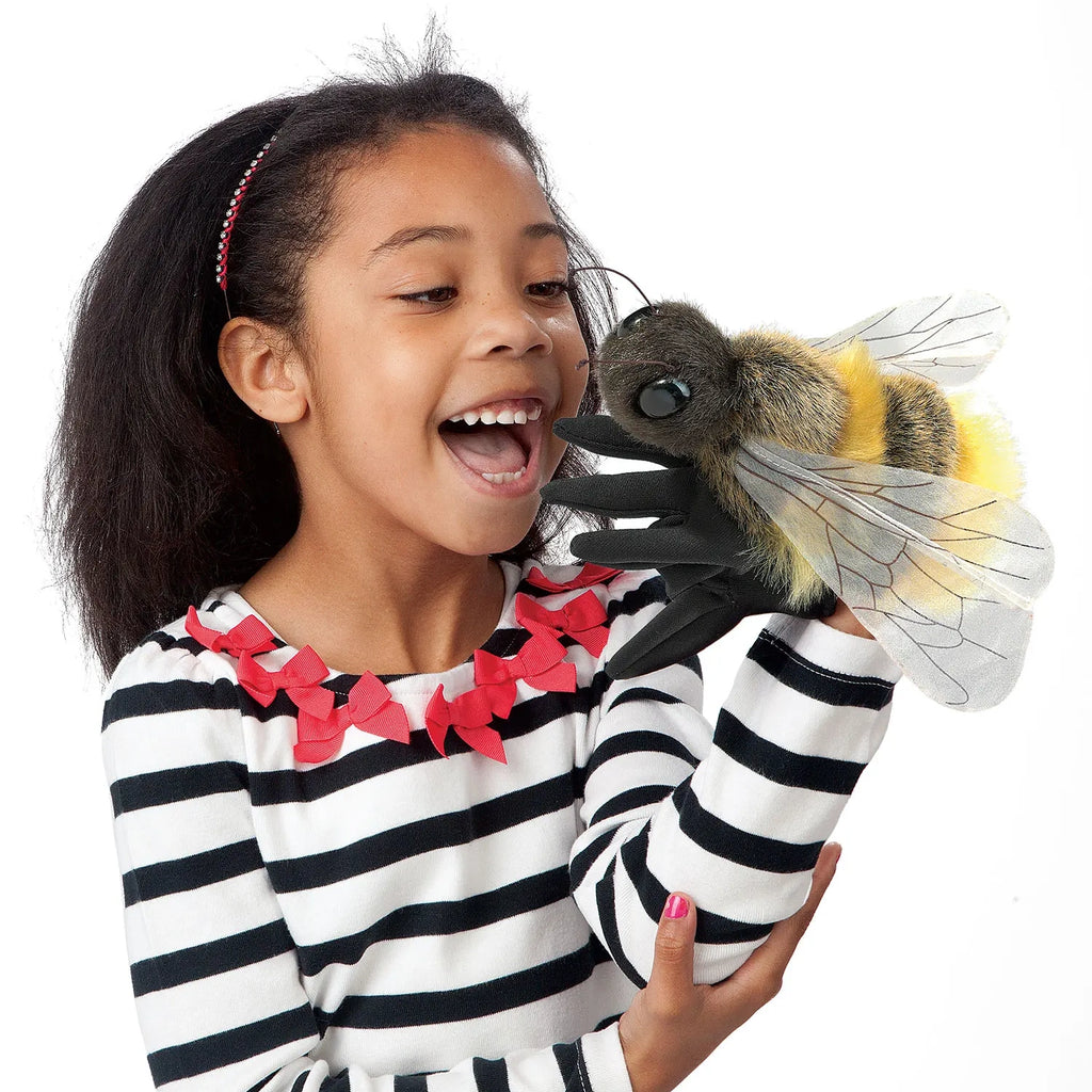 A young girl in a striped shirt joyfully interacts with a large bee puppet on her hand, smiling brightly, showing excitement and wonder.