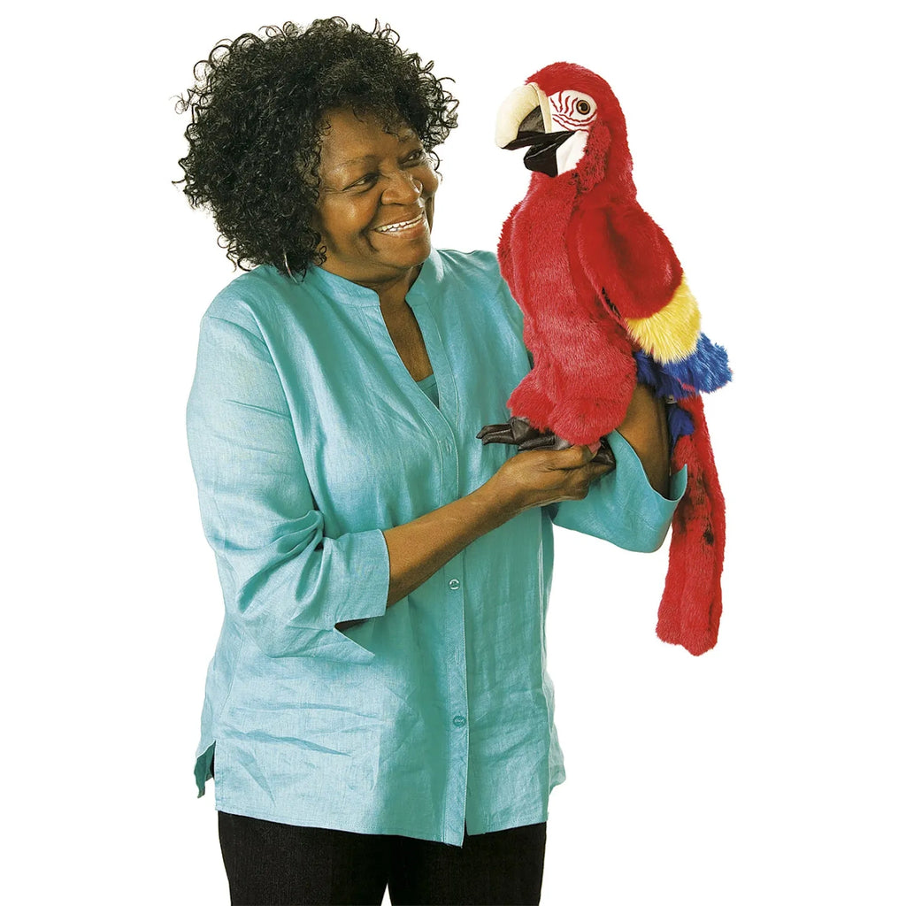 A smiling woman in a turquoise shirt holds a vibrant toy parrot. The parrot is red with yellow and blue wings, creating a joyful and playful scene.