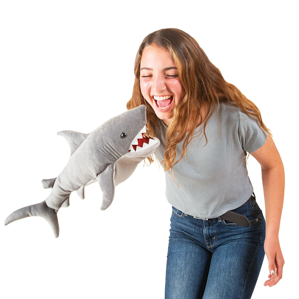A woman in jeans and a gray shirt laughs joyfully while playfully holding a plush shark. The shark puppet appears to be "biting" her arm.