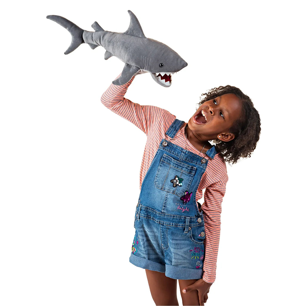 A joyful child in striped shirt and denim overalls holds a plush shark above her head, pretending to roar. She exudes excitement and playfulness.
