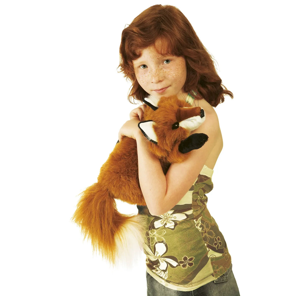 A child with red hair and freckles lovingly hugs a plush red fox. She wears a floral, green top and jeans, creating a warm and playful atmosphere.