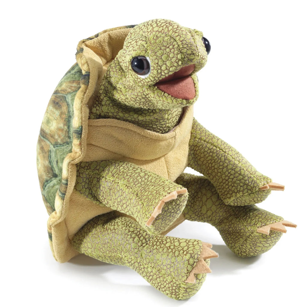 A plush tortoise puppet with textured green skin and big, friendly eyes sits upright. Its open mouth gives a cheerful expression.