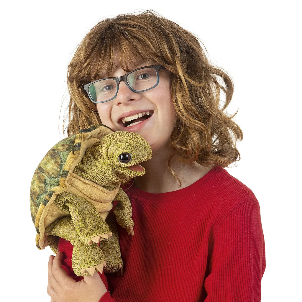 A smiling child with long, wavy hair and glasses holds a realistic turtle puppet. The child wears a red sweater, creating a joyful and playful atmosphere.