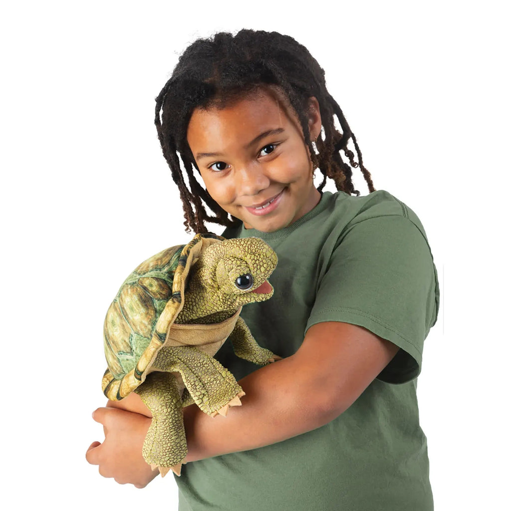 A child with curly hair and a green shirt smiles warmly, holding a plush turtle puppet. The scene is bright, conveying joy and playfulness.