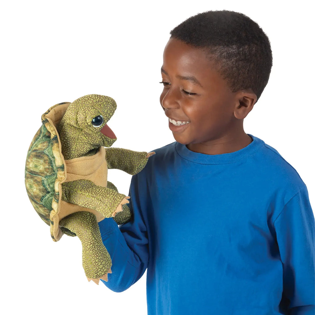A smiling boy in a blue shirt holds a green turtle puppet on his hand, looking at it with joy. The puppet is detailed with textured skin.