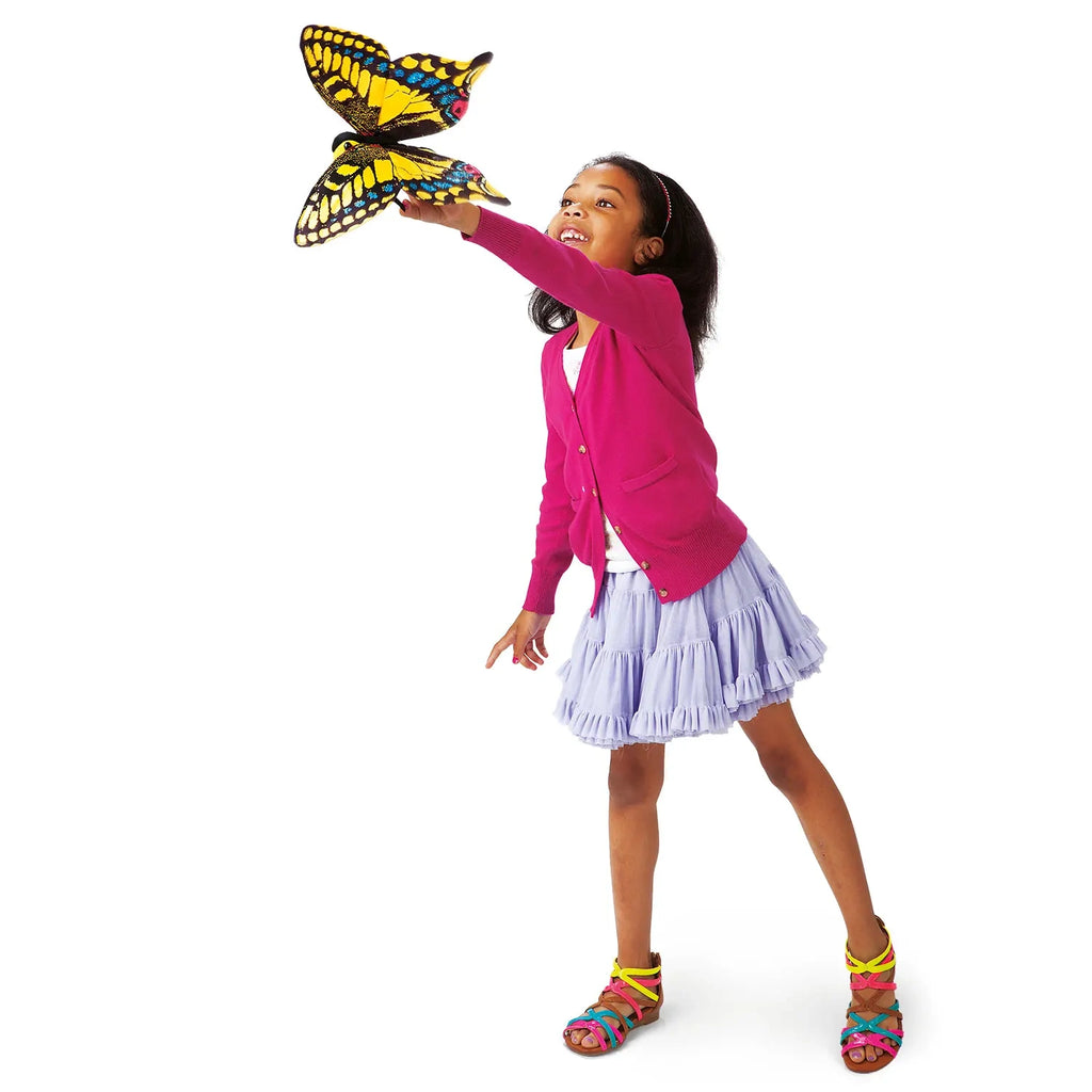 A joyful child in a pink sweater and purple skirt reaches toward a vibrant yellow and black butterfly in mid-flight, expressing wonder and delight.