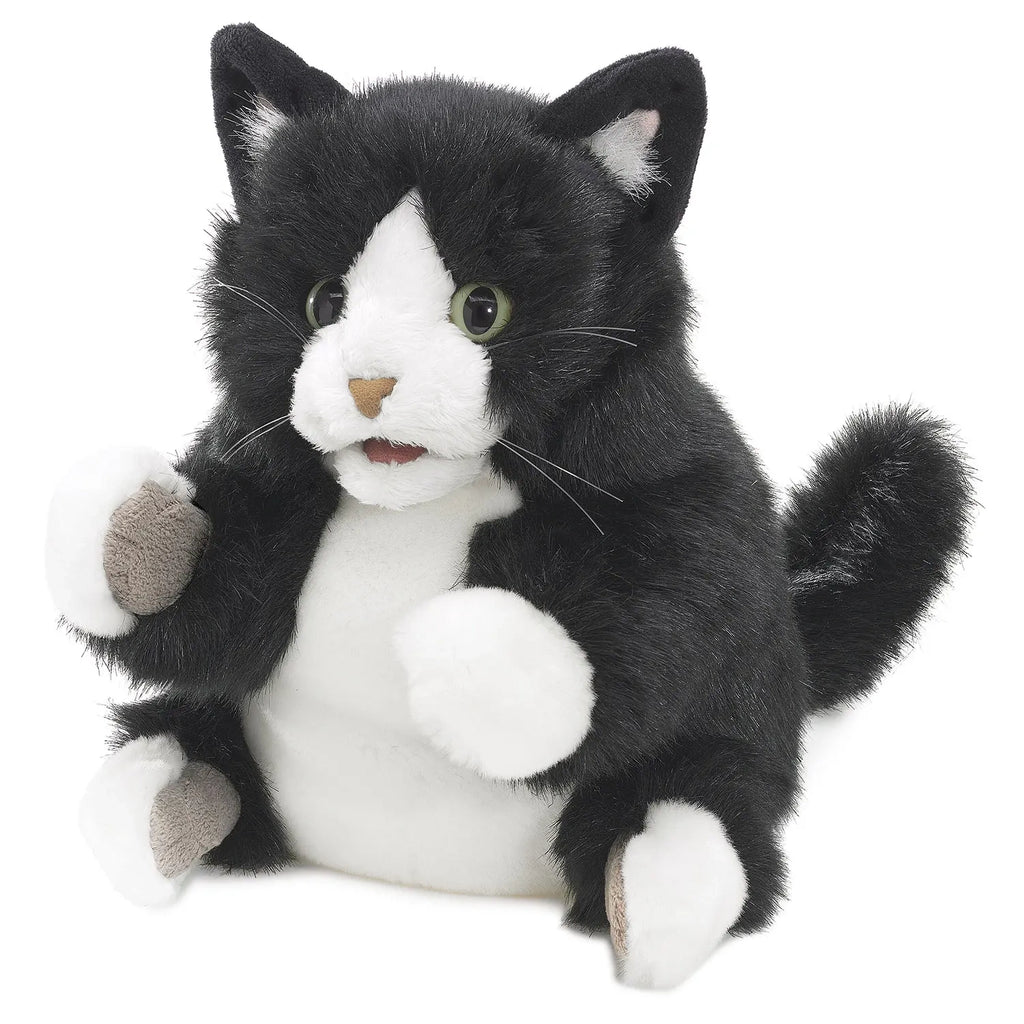 A plush toy cat with black and white fur sits upright. It has wide eyes and a soft, friendly expression, conveying a playful and cuddly vibe.