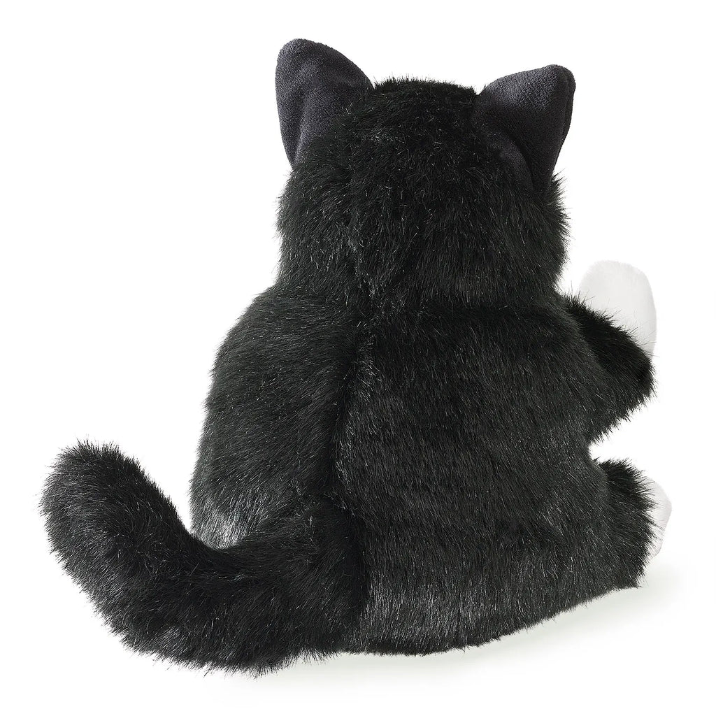 A fluffy black and white plush cat is seen from behind. It sits with an upright tail and ears pointed forward, conveying a playful and curious tone.