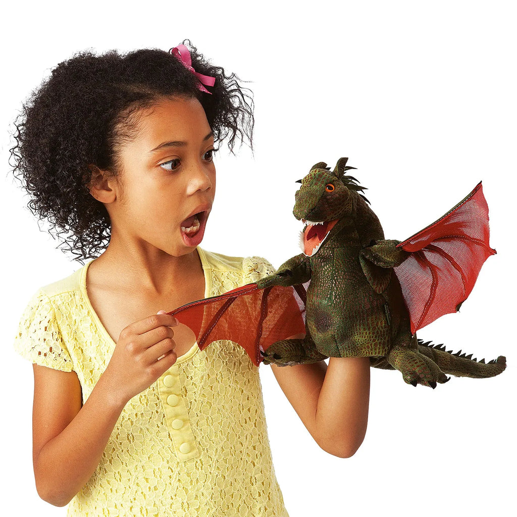 A surprised young girl in a yellow dress holds a dragon puppet with green skin and red wings. The playful scene conveys excitement and wonder.