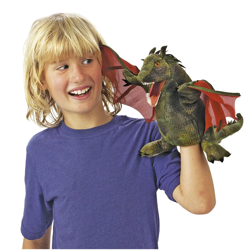 A smiling child in a blue shirt holds a green dragon puppet with red wings on their hand. The playful scene conveys joy and imagination.