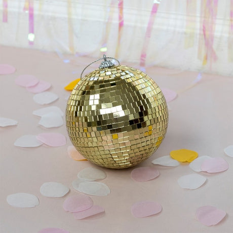 A single gold disco ball sits on a light pink surface surrounded by scattered pastel confetti.
