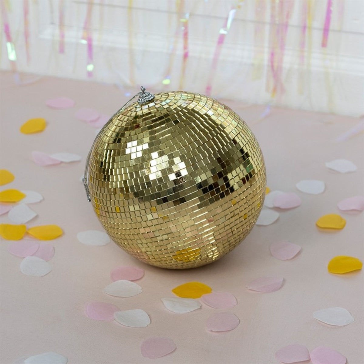 A shiny gold disco ball rests on a pink surface scattered with white, pink, and yellow confetti.