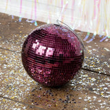 A plum purple disco ball with a silver hanging loop sits on a wooden floor covered in scattered star confetti.
