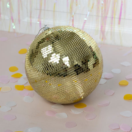 A large gold disco ball resting on a light pink surface surrounded by yellow and white paper confetti.