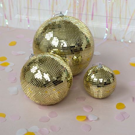 Three shimmering gold disco balls of different sizes are arranged on a light pink surface scattered with yellow, white, and pink confetti.