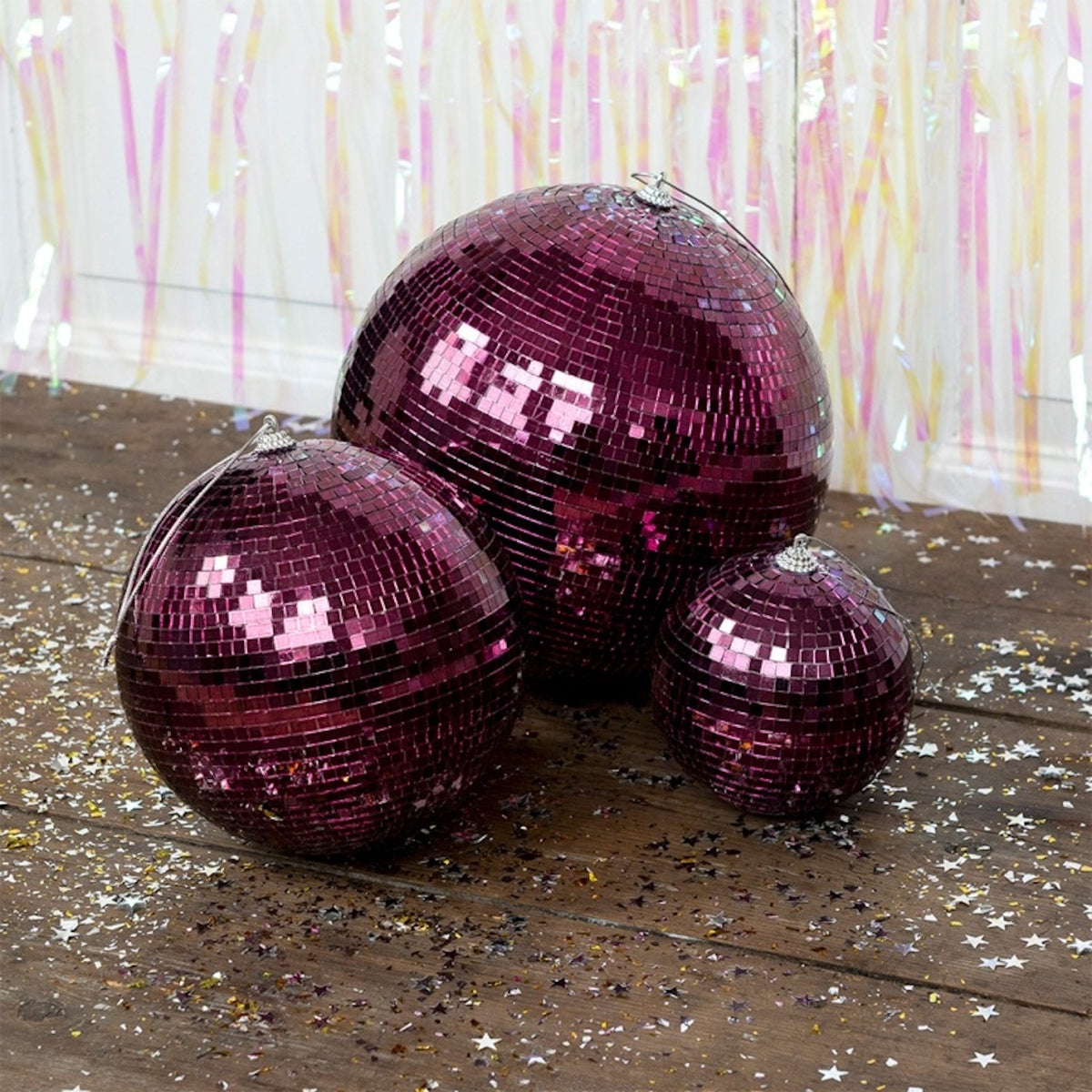 Three plum-purple disco balls of varying sizes sit on a wooden floor scattered with star confetti.