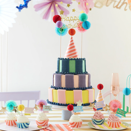 Three-tiered birthday cake topped with a striped hat and a gold "Happy Birthday" topper surrounded by cupcakes.