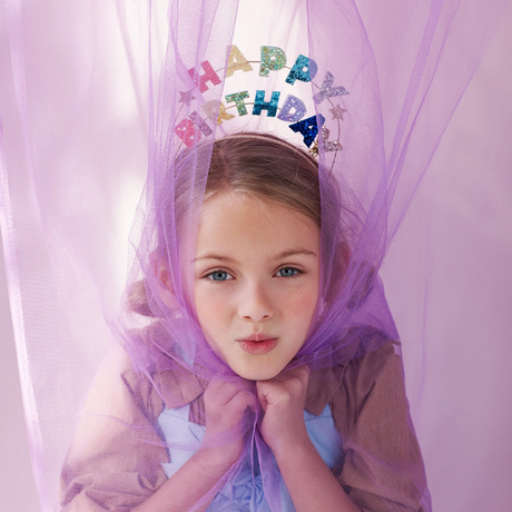 Young girl in a purple tulle veil wears a glittery “HAPPY BIRTHDAY” crown, posing with hands under her chin in a soft, playful portrait.