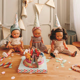 Three baby dolls wearing party hats sitting at a wooden table with a wooden toy birthday cake and confetti.