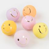 Six pastel-colored paper surprise balls wrapped with shiny gold smiley face faces on a white background.