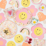 Flat lay view of a colorful Meri Meri party spread featuring smiley flower plates, patterned napkins, and various 3D honeycomb cups.