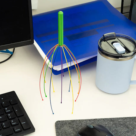 A colorful head massager with a green handle stands upright on a desk near a keyboard, blue folder, light blue travel mug, and computer mouse.