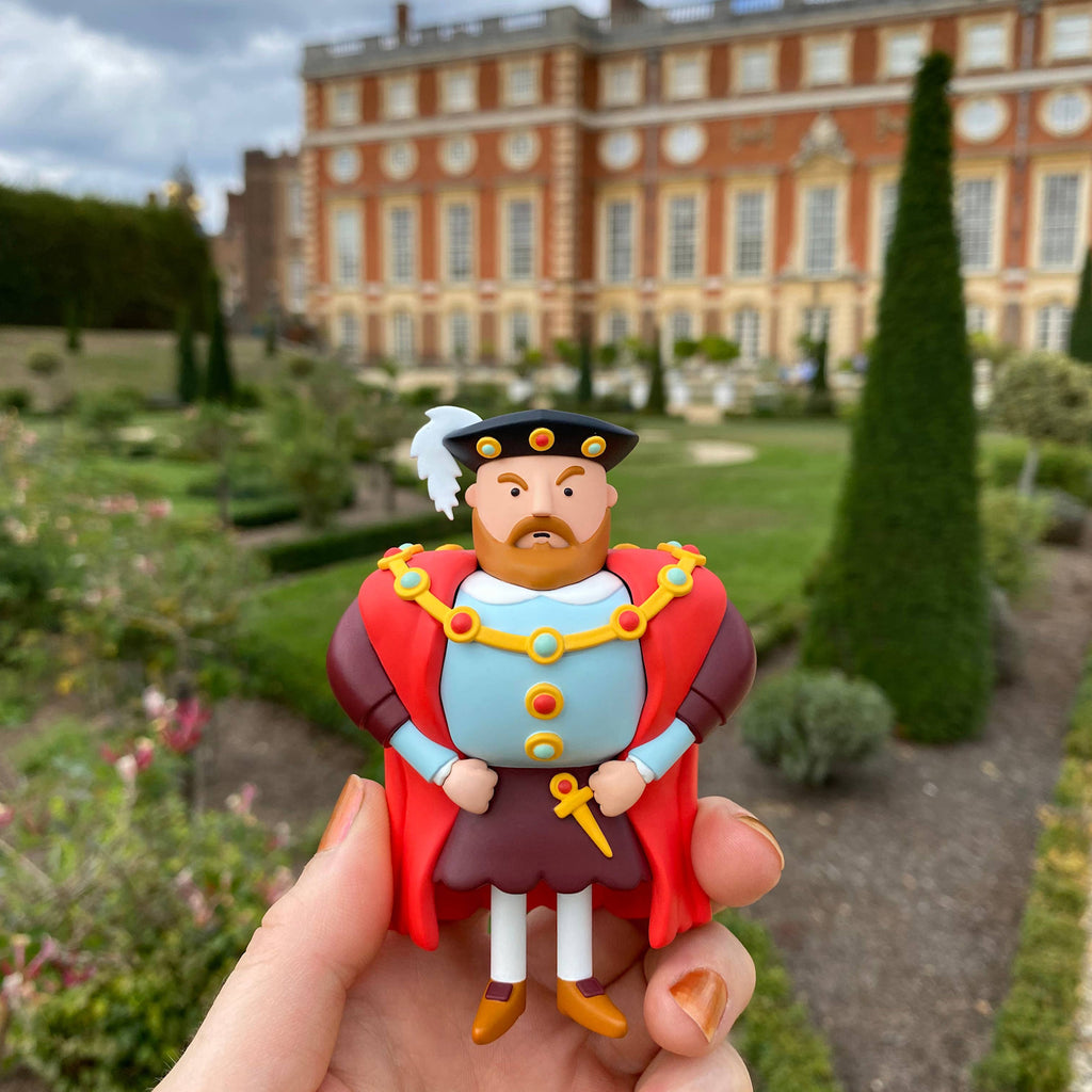 A small, colorful toy figure of a regal character is held up in front of an elegant historic manor with manicured gardens, exuding charm and whimsy.