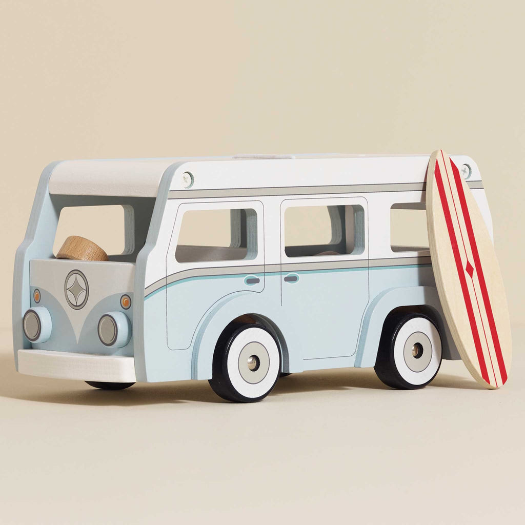 Toy vintage van in light blue with white details, featuring a detachable roof. A stylish surfboard with red stripes leans against the van, evoking a fun, coastal vibe.