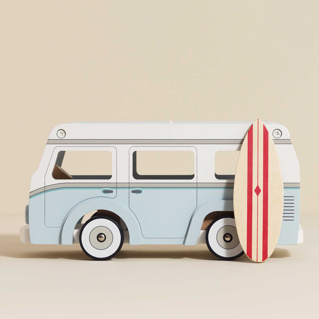 Toy vintage van in light blue with white details, featuring a detachable roof. A stylish surfboard with red stripes leans against the van, evoking a fun, coastal vibe.