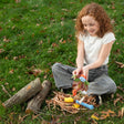 A young girl with curly hair sits on grass, crafting colorful clay creatures with leaves and twigs in a straw nest.