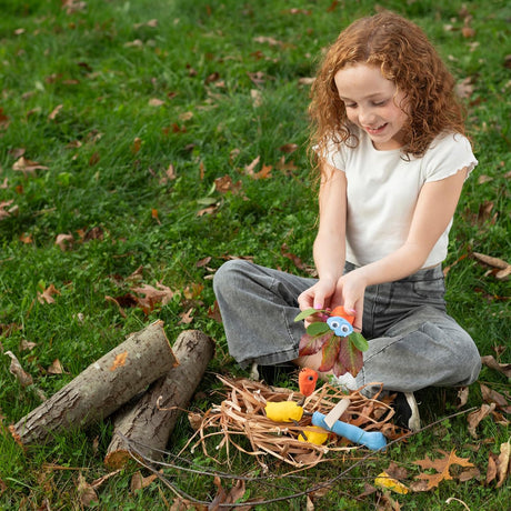 A young girl with curly hair sits on grass, crafting colorful clay creatures with leaves and twigs in a straw nest.