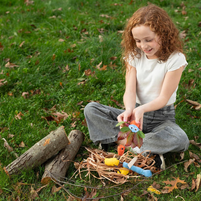 A young girl with curly hair sits on grass, crafting colorful clay creatures with leaves and twigs in a straw nest.