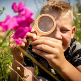 A child using a wooden Huckleberry dual magnifier to inspect vibrant pink flowers in a sunny outdoor field.