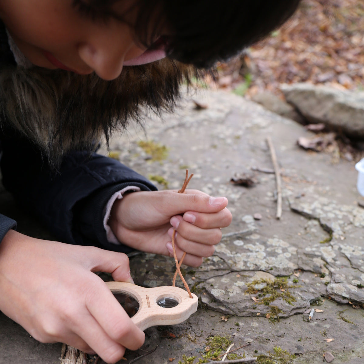 Child using a Huckleberry wooden dual magnifier to explore moss and rocks outdoors, nature study and educational toy.