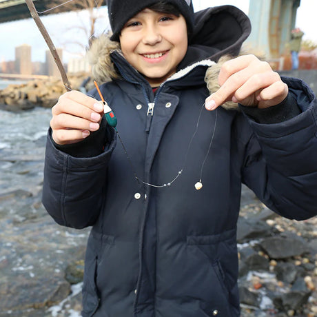 Smiling child in a winter coat holding a stick fishing pole with a bobber and line by a rocky shoreline.