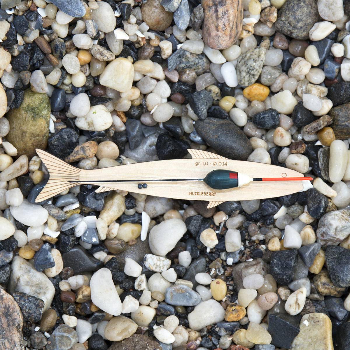 A wooden fish-shaped hand line with a float and fishing line sits on a bed of smooth river stones and pebbles.