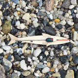 A wooden fish-shaped hand line with a float and fishing line sits on a bed of smooth river stones and pebbles.