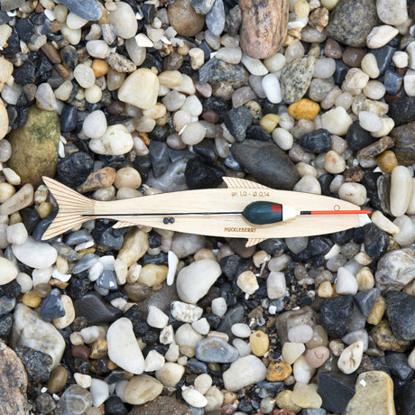 A wooden fish-shaped hand line with a float and fishing line sits on a bed of smooth river stones and pebbles.