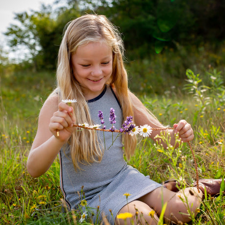 Young girl in a meadow smiling while crafting a DIY necklace with fresh daisies and lavender on a braided cord.