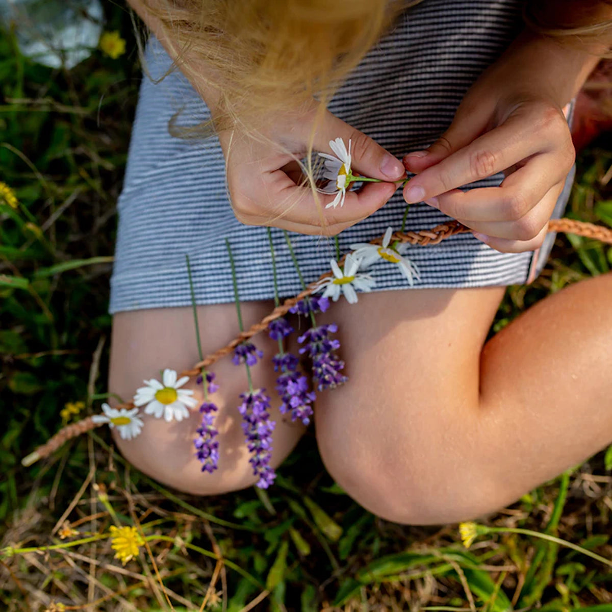 A child weaving fresh daisies and lavender into a braided tan necklace cord for a DIY nature craft activity.