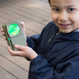 Young boy holding a green Huckleberry Morse code signal light toy outdoors on a wooden deck, showing a glowing green lens.