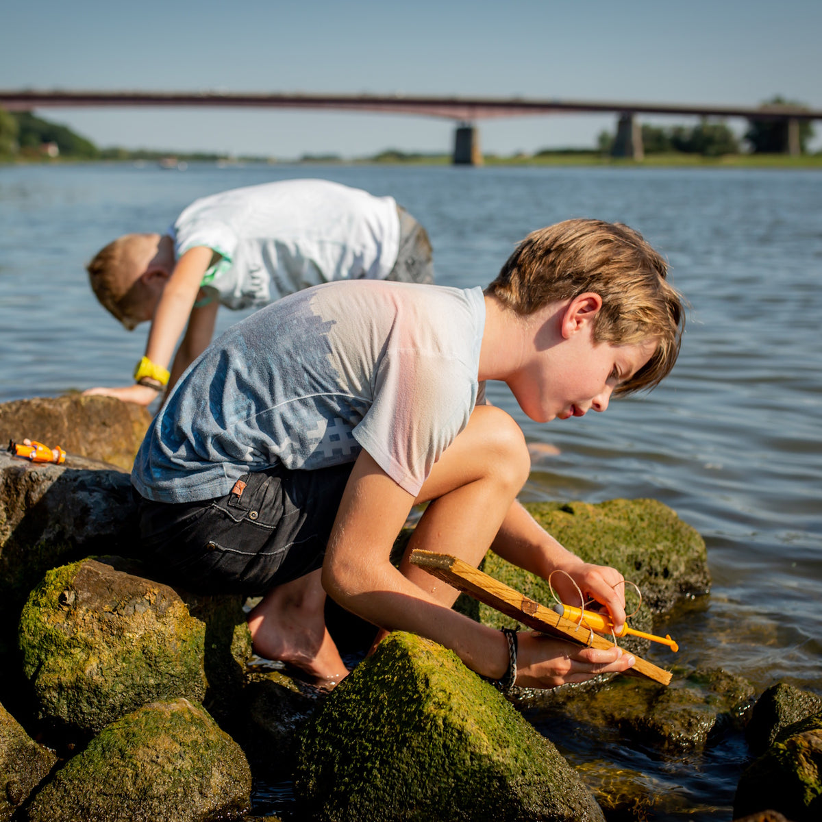 Two boys playing with a toy motor boat on mossy river rocks with a bridge in the background on a sunny day.
