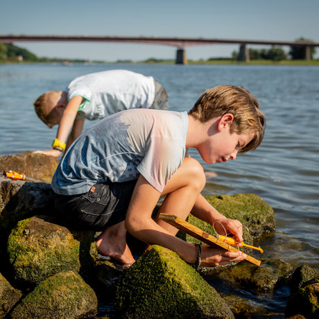 Two boys playing with a toy motor boat on mossy river rocks with a bridge in the background on a sunny day.