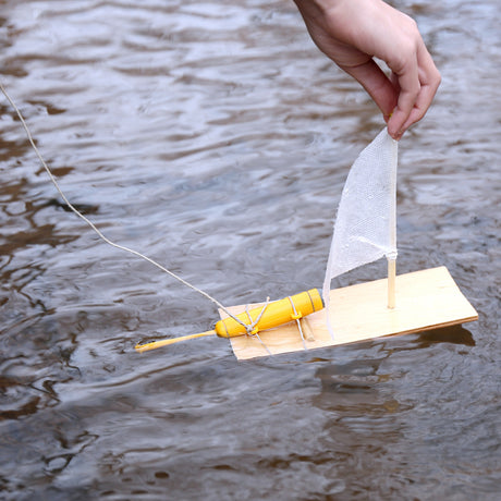 Small wooden toy motor boat with a yellow motor and white sail floating on water, being held by a hand.