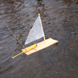 Wooden Huckleberry motor boat toy with white sail and yellow propeller motor floating on dark rippling water.