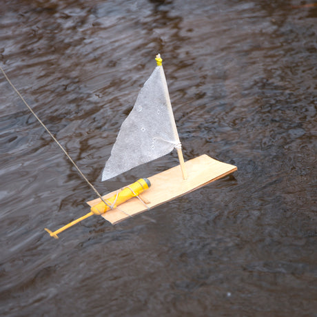 Wooden Huckleberry motor boat toy with white sail and yellow propeller motor floating on dark rippling water.