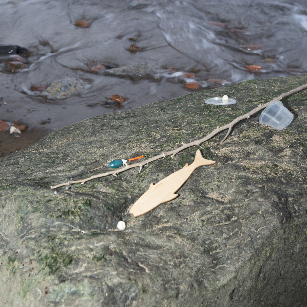 A wooden fish-shaped hand reel and fishing line with a bobber rest on a rock by a stream.